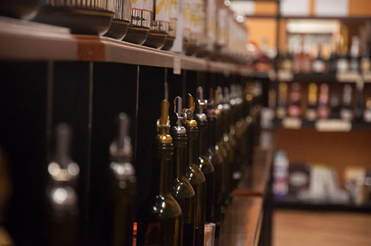 A row of dark glass olive oil bottles displayed at a specialty store, with open samples available for customers to smell and taste, demonstrating proper light-protective packaging and the retailer's transparency in allowing quality evaluation before purchase.