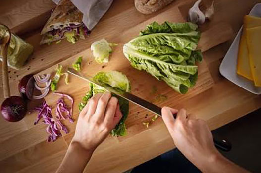 Overhead view of hands slicing romaine lettuce on a wooden cutting board with fresh vegetables nearby